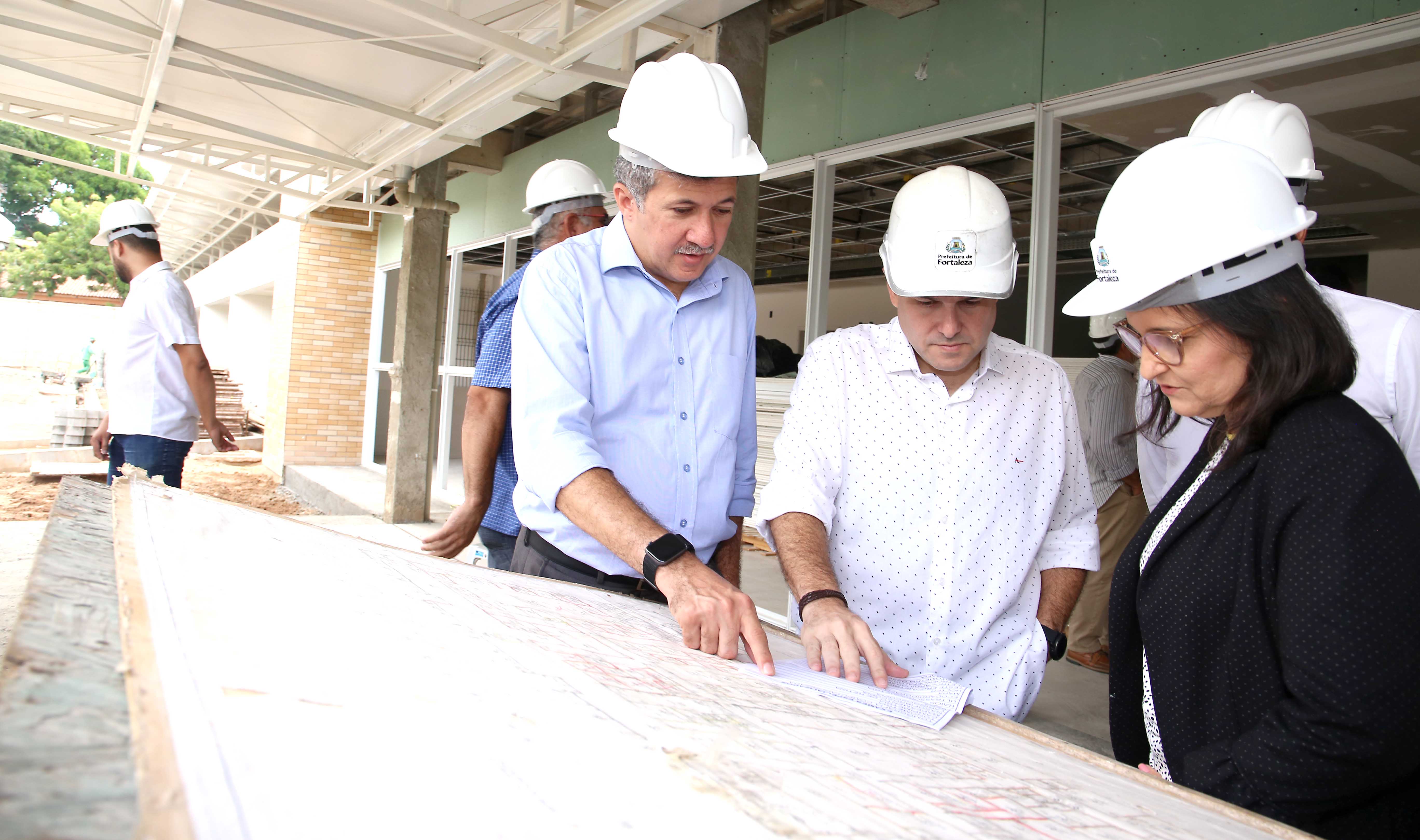 prefeito e pessoas ao lado com capacete de engenheiro analisando papel em cima de mesa